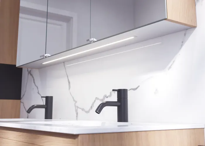 Badkamerinspiratie met wastafelkasten en bovenkasten in licht eikenhout met groeven, elegante dubbele wastafel, mooie spiegelkast met verlichting, visgraatvloer in licht hout en witte marmeren muur geven een klassieke, lichte en strakke uitstraling.