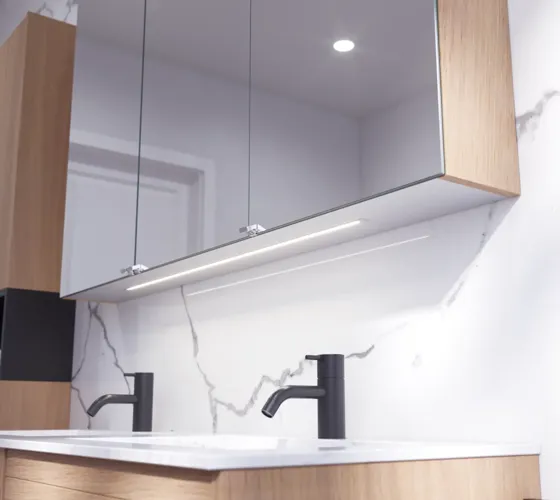 Badkamerinspiratie met wastafelkasten en bovenkasten in licht eikenhout met groeven, elegante dubbele wastafel, mooie spiegelkast met verlichting, visgraatvloer in licht hout en witte marmeren muur geven een klassieke, lichte en strakke uitstraling.