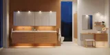 Moderne badkamer in Noordse stijl met licht hout, elegante verlichting, spiegelkastmuur en stijlvolle inrichting die een warme en uitnodigende sfeer creëert.