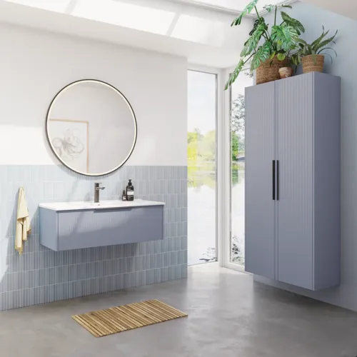 Badkamerinspiratie met lichtblauwe tegels en betonnen vloer, lichtblauw wastafelmeubel met smalle strepen, dunne wastafel, dubbele hoge kast en een ronde spiegel met zwarte rand en verlichting.